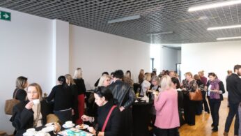 Ujęcie z przerwy kawowej lub lunchu, na którym duża grupa osób stoi i rozmawia przy stolikach. Uczestnicy jedzą i piją, korzystając z czasu na networking. Wnętrze charakteryzuje się jasnymi ścianami i kratownicowym sufitem.