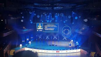 Szerokie ujęcie z góry na dużą salę widowiskową z pełną widownią i rozbudowaną scenografią. Na scenie, oświetlonej na niebiesko, występuje prelegent, a za nim widoczny jest duży ekran z prezentacją, która zawiera listę punktów. Widoczne są logo sponsorów wydarzenia.