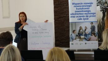Kobieta z rudymi włosami prezentująca planszę z tekstem podczas spotkania, obok niej stoi duży baner promocyjny.