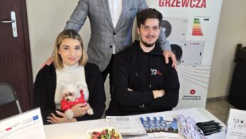 Trzy osoby, dwóch mężczyzn i kobieta, stoją za stołem z materiałami promocyjnymi firmy ND Polska, zajmującej się techniką grzewczą. Kobieta trzyma w ramionach małego, białego psa. Na stole leżą słodycze, notesy, przypinki i smycze.