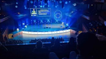 Szerokie ujęcie na dużą scenę w nowoczesnej sali widowiskowej podczas prezentacji, wykonane z tylnych rzędów widowni. Na scenie występuje prelegent, a na ekranie widoczne są slajdy w języku angielskim. Scenografia jest oświetlona na niebiesko i zawiera geometryczne wzory.