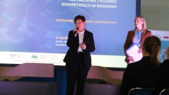 Dwie kobiety stoją na scenie przed dużym ekranem z napisem 