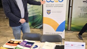 Mężczyzna w okularach i marynarce stoi obok stoiska Opolskeigo Centrum Rozwoju Gospodarki, wskazując ręką na baner. Na stole leży wiele materiałów promocyjnych, w tym broszury o różnych branżach, notesy i ołówki.