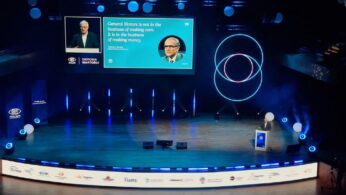 Szerokie ujęcie sceny w dużej, nowoczesnej sali konferencyjnej, oświetlonej na niebiesko. Na scenie stoi prelegent, a za nim znajduje się duży ekran z prezentacją. Ekran wyświetla cytat w języku angielskim przypisywany Thomasowi A. Murphy'emu z General Motors, dotyczący biznesu i zarabiania pieniędzy. Na dole sceny widoczne są logotypy sponsorów wydarzenia 