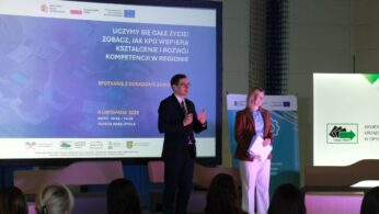 Dwóch prelegentów stoi na scenie przed dużym ekranem. Mężczyzna w garniturze i krawacie mówi do mikrofonu, gestykulując. Obok niego stoi kobieta w jasnym żakiecie, trzymająca clipboard. Na ekranie wyświetlony jest napis: 
