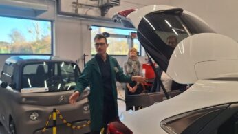 Mężczyzna w zielonym fartuchu stoi między dwoma samochodami: szarym, małym Citroenem Ami i białym samochodem z otwartym bagażnikiem. Mężczyzna gestykuluje, prezentując wnętrze pojazdu. W tle widać osoby obserwujące prezentację w nowoczesnym warsztacie.
