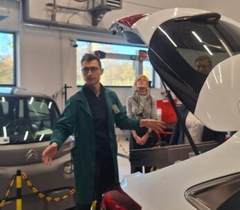 Mężczyzna w zielonym fartuchu stoi między dwoma samochodami: szarym, małym Citroenem Ami i białym samochodem z otwartym bagażnikiem. Mężczyzna gestykuluje, prezentując wnętrze pojazdu. W tle widać osoby obserwujące prezentację w nowoczesnym warsztacie.