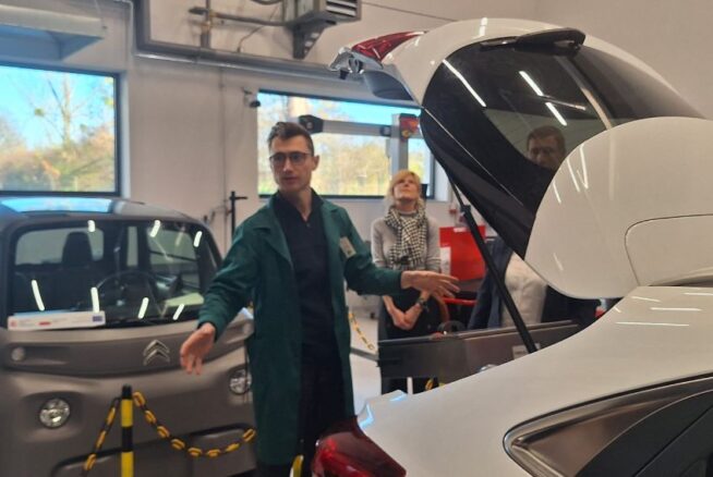Mężczyzna w zielonym fartuchu stoi między dwoma samochodami: szarym, małym Citroenem Ami i białym samochodem z otwartym bagażnikiem. Mężczyzna gestykuluje, prezentując wnętrze pojazdu. W tle widać osoby obserwujące prezentację w nowoczesnym warsztacie.