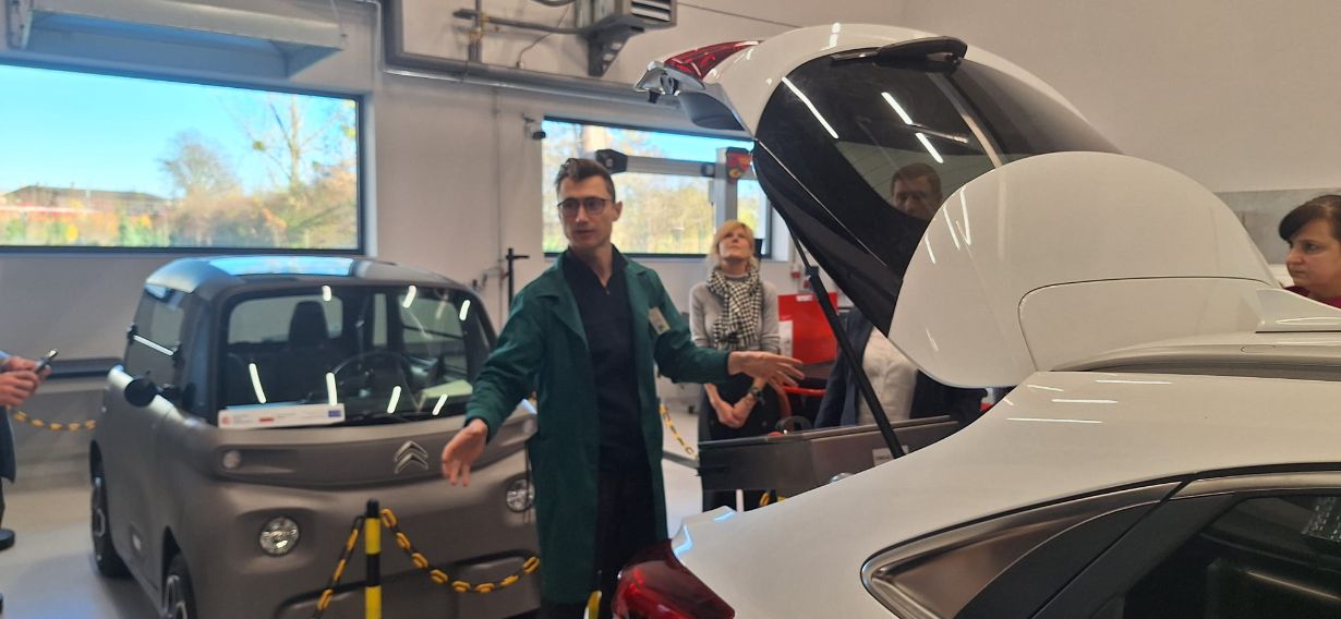 Mężczyzna w zielonym fartuchu stoi między dwoma samochodami: szarym, małym Citroenem Ami i białym samochodem z otwartym bagażnikiem. Mężczyzna gestykuluje, prezentując wnętrze pojazdu. W tle widać osoby obserwujące prezentację w nowoczesnym warsztacie.