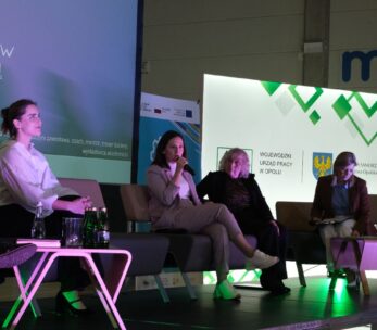 Cztery kobiety siedzą na kanapach na scenie podczas panelu dyskusyjnego, z widocznymi elementami brandingowymi Urzędu Pracy i Samorządu Województwa Opolskiego. Jedna z pań trzyma mikrofon i aktywnie uczestniczy w dyskusji. Obok stolików leżą butelki z wodą.