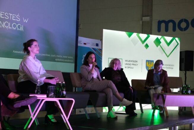 Cztery kobiety siedzą na kanapach na scenie podczas panelu dyskusyjnego, z widocznymi elementami brandingowymi Urzędu Pracy i Samorządu Województwa Opolskiego. Jedna z pań trzyma mikrofon i aktywnie uczestniczy w dyskusji. Obok stolików leżą butelki z wodą.