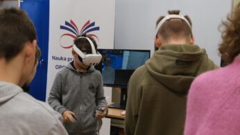 Chłopiec w szarej bluzie z kapturem ma na głowie gogle VR i trzyma kontrolery, skupiony na wirtualnym doświadczeniu. Stoi przed monitorem, na którym widać wnętrze warsztatu z narzędziami. W tle widoczny jest baner z hasłem 
