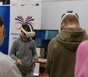 Chłopiec w szarej bluzie z kapturem ma na głowie gogle VR i trzyma kontrolery, skupiony na wirtualnym doświadczeniu. Stoi przed monitorem, na którym widać wnętrze warsztatu z narzędziami. W tle widoczny jest baner z hasłem "Nauka przez całe życie" oraz fragmenty sylwetek innych osób.
