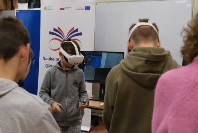 Chłopiec w szarej bluzie z kapturem ma na głowie gogle VR i trzyma kontrolery, skupiony na wirtualnym doświadczeniu. Stoi przed monitorem, na którym widać wnętrze warsztatu z narzędziami. W tle widoczny jest baner z hasłem "Nauka przez całe życie" oraz fragmenty sylwetek innych osób.