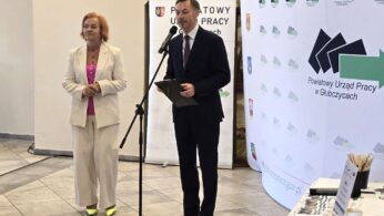 Kobieta w jasnym garniturze i mężczyzna w ciemnym garniturze stoją przy mikrofonie, wygłaszając przemówienie. Za nimi znajduje się duży baner Powiatowego Urzędu Pracy w Głubczycach, zawierający też logotypy programów finansowanych przez Unię Europejską. Jest to ujęcie z uroczystości otwarcia.