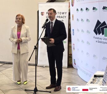 Kobieta w jasnym garniturze i mężczyzna w ciemnym garniturze stoją przy mikrofonie, wygłaszając przemówienie. Za nimi znajduje się duży baner Powiatowego Urzędu Pracy w Głubczycach, zawierający też logotypy programów finansowanych przez Unię Europejską. Jest to ujęcie z uroczystości otwarcia.