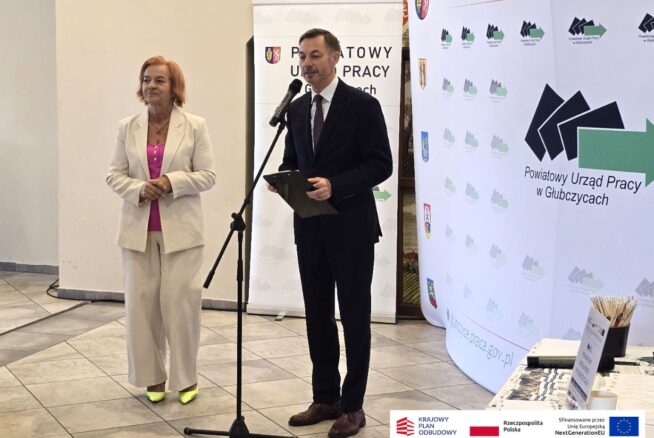Kobieta w jasnym garniturze i mężczyzna w ciemnym garniturze stoją przy mikrofonie, wygłaszając przemówienie. Za nimi znajduje się duży baner Powiatowego Urzędu Pracy w Głubczycach, zawierający też logotypy programów finansowanych przez Unię Europejską. Jest to ujęcie z uroczystości otwarcia.
