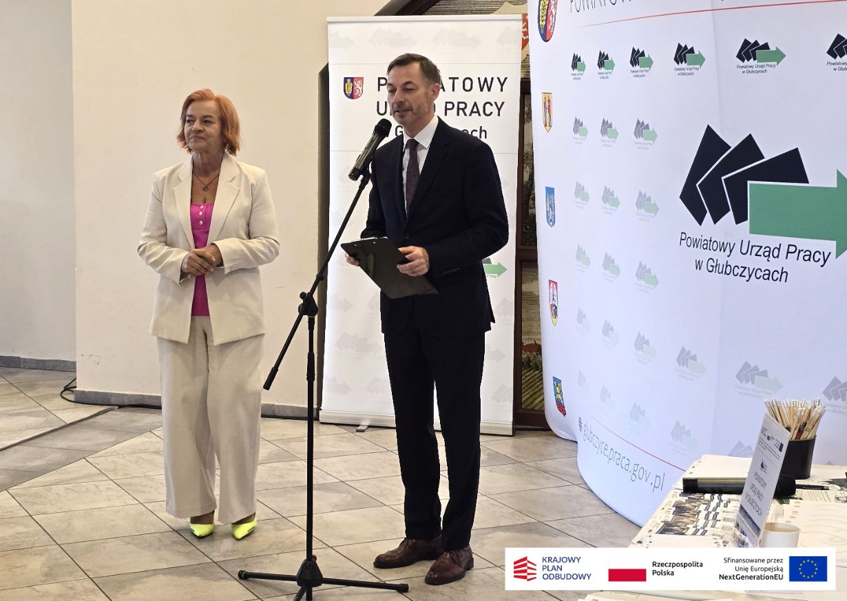 Kobieta w jasnym garniturze i mężczyzna w ciemnym garniturze stoją przy mikrofonie, wygłaszając przemówienie. Za nimi znajduje się duży baner Powiatowego Urzędu Pracy w Głubczycach, zawierający też logotypy programów finansowanych przez Unię Europejską. Jest to ujęcie z uroczystości otwarcia.