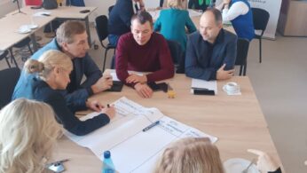 Uczestnicy warsztatów siedzą wokół stołu, aktywnie pracując nad zadaniem. Jedna z kobiet notuje na dużym arkuszu papieru. W tle widoczne są flagi Polski i Unii Europejskiej oraz banery promocyjne, w tym jeden z nazwiskiem wicemarszałek Zuzanny Donath-Kasiury.