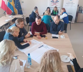 Uczestnicy warsztatów siedzą wokół stołu, aktywnie pracując nad zadaniem. Jedna z kobiet notuje na dużym arkuszu papieru. W tle widoczne są flagi Polski i Unii Europejskiej oraz banery promocyjne, w tym jeden z nazwiskiem wicemarszałek Zuzanny Donath-Kasiury.