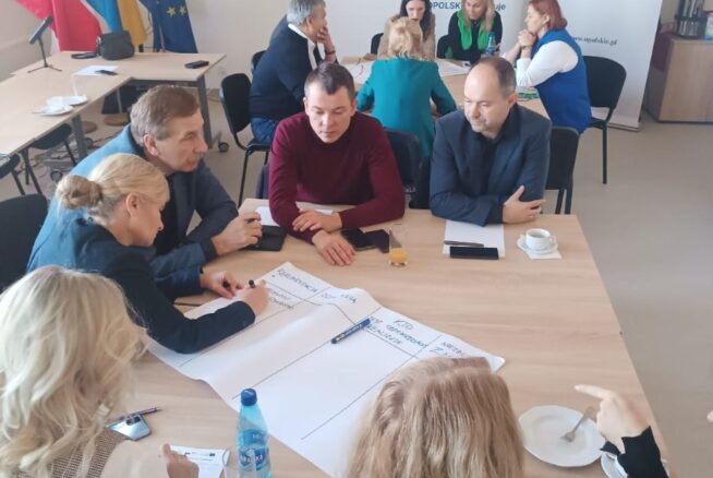 Uczestnicy warsztatów siedzą wokół stołu, aktywnie pracując nad zadaniem. Jedna z kobiet notuje na dużym arkuszu papieru. W tle widoczne są flagi Polski i Unii Europejskiej oraz banery promocyjne, w tym jeden z nazwiskiem wicemarszałek Zuzanny Donath-Kasiury.