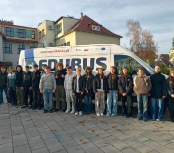 Grupa młodzieży stoi przed białym busem z napisem "EDUBUS" i adresem internetowym "www.opolskieLLL.pl". Bus jest zaparkowany na brukowanym placu z jesiennymi liśćmi. W tle widać budynki szkolne i wieżę kościoła.