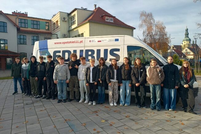 Grupa młodzieży stoi przed białym busem z napisem "EDUBUS" i adresem internetowym "www.opolskieLLL.pl". Bus jest zaparkowany na brukowanym placu z jesiennymi liśćmi. W tle widać budynki szkolne i wieżę kościoła.
