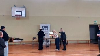 Na zdjęciu widać dwie młode uczennice szkoły podstawowej w sali gimnastycznej, korzystające z gogli VR. Stoją w środku pomieszczenia, wykonując ruchy kontrolerami. W tle znajdują się dwa monitory prezentujące widok z gogli VR oraz roll-up projektu, promujący „EduBusa – mobilny punkt szerzenia idei Lifelong Learning” na Opolszczyźnie. Nad nimi widoczny jest kosz do koszykówki i wysokie okna oświetlające salę. Uczennice otoczone są kolegami z klasy obserwującymi ich działania w aplikacji „Widzę siebie w tym zawodzie” na monitorach.
