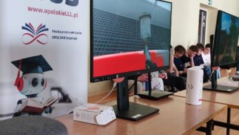 Widok na dwie szkolne ławki ustawione w Sali gimnastycznej w szkole podstawowej. Na ławkach znajdują się dwa monitory komputerowe oraz futerał od zestawu Gogli VR, które podłączone są do monitorów. W tle, za ławkami roll-up promujący „EduBus”, a obok niego grupa uczniów czekająca na swoją kolej do skorzystania z gogli VR.