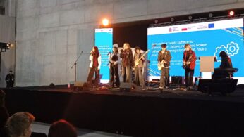 Duża scena w przestronnej hali, w której naprzeciwko sceny znajdują się miejsca siedzące wypełnione młodą publiką. Na scenie znajduje się grupa – zespół młodych muzyków wykonujących utwór wokalnie, z użyciem instrumentów (gitar). Za wykonawcami muzycznymi znajduje się duża ściana reklamowa promująca projekt 