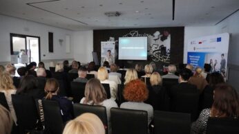 Szeroki kadr przedstawiający dużą grupę osób siedzącą w rzędach krzeseł, uczestniczącą w szkoleniu lub konferencji. Na scenie, przed dużym ekranem z widocznym slajdem, stoi prelegent. Z tyłu sali widoczny jest banner promujący rozwój nowoczesnego szkolnictwa zawodowego w Powiecie Nyskim.
