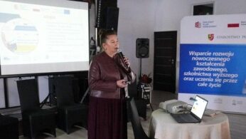 Zdjęcie przestawia kobietę – prelegentkę przemawiającą do trzymanego w dłoni mikrofonu. Za kobietą znajduje się roll-up promujący projekt „Wsparcie rozwoju nowoczesnego kształcenia zawodowego, szkolnictwa wyższego oraz uczenia się przez cale życie”, a obok niej fragment ekranu, na którym wyświetlana jest prezentacja z treścią wystąpienia.
