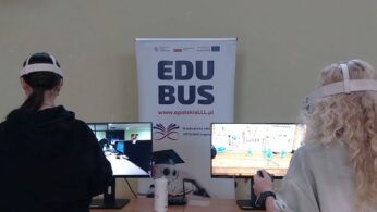 widok zza pleców dwóch młodych uczennic szkoły podstawowej, które znajdują się w sali gimnastycznej, korzystających z gogli VR. Uczennice z zaangażowaniem wykonują ruchy kontrolerami. W tle znajdują się dwa monitory prezentujące widok z gogli VR oraz roll-up projektu, promujący „EduBusa – mobilny punkt szerzenia idei Lifelong Learning” na Opolszczyźnie.