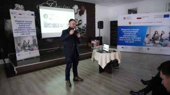 Zdjęcie prelegenta stojącego na scenie, ubranego w marynarkę i dżinsy, trzymającego mikrofon w dłoni. W tle widoczny jest ekran z nazwiskiem 