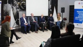 Zdjęcie przestawia uczestników spotkania biorących żywy udział w debacie. Dyskutują, zabierają głos do mikrofonu. Wszyscy znajdują się dość przestronnej sali zajmując miejsca obok siebie jak w panelu dyskusyjnym. Z boku oraz naprzeciw nich reszta uczestników spotkania. Otoczeni są roll-upem oraz ścianką reklamową, promującymi projekt „Wsparcie rozwoju nowoczesnego kształcenia zawodowego, szkolnictwa wyższego oraz uczenia się przez cale życie”, a obok niej fragment ekranu, na którym wyświetlana jest prezentacja z treścią wystąpienia.