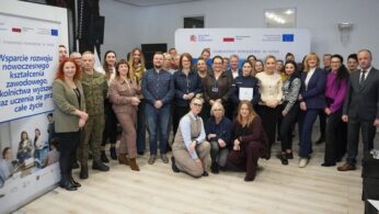 Zdjęcie grupowe, przedstawiające wszystkich zgromadzonych uczestników spotkania roboczego ustawionych do wspólnej fotografii. Wszyscy znajdują się dość przestronnej, jasno oświetlonej Sali, zajmując miejsca obok siebie tworząc dwa rzędy postaci. W tylnym rzędzie większość uczestników, razem 30 osób i trzy osoby w „przykucu”, tworzące mały rząd frontowy. Otoczeni są roll-upem oraz ścianką reklamową, promującymi projekt „Wsparcie rozwoju nowoczesnego kształcenia zawodowego, szkolnictwa wyższego oraz uczenia się przez cale życie”.