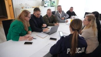 Grupa osób, w tym cywile, wojskowy i policjantka w mundurze (tyłem do obiektywu), siedzi przy dużym białym stole podczas spotkania roboczego. Mężczyzna w centrum wskazuje coś na ekranie laptopa, a reszta aktywnie uczestniczy w dyskusji. Spotkanie to prawdopodobnie dotyczy współpracy w ramach projektu edukacyjnego.