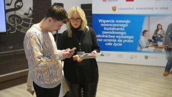 Dwie osoby stoją na tle banneru promującego rozwój kształcenia zawodowego. Mężczyzna w kolorowym swetrze patrzy na swój telefon komórkowy, a kobieta w okularach i czarnym golfie trzyma w rękach folder z dokumentami. Jest to moment przerwy lub wymiany informacji podczas konferencji.