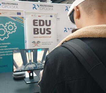 Ujęcie od tyłu, młodego ucznia z założonymi goglami VR. Uczeń skierowany jest w stronę ekranu, na którym wyświetla się kadr z aplikacji „Widzę siebie w tym zawodzie”. W tle, za ekranem znajduje się banner promujący projekt "Wsparcie rozwoju nowoczesnego kształcenia zawodowego..." oraz banner promujący „EduBusa – mobilny punkt szerzenia idei Lifelong Learning” na Opolszczyźnie