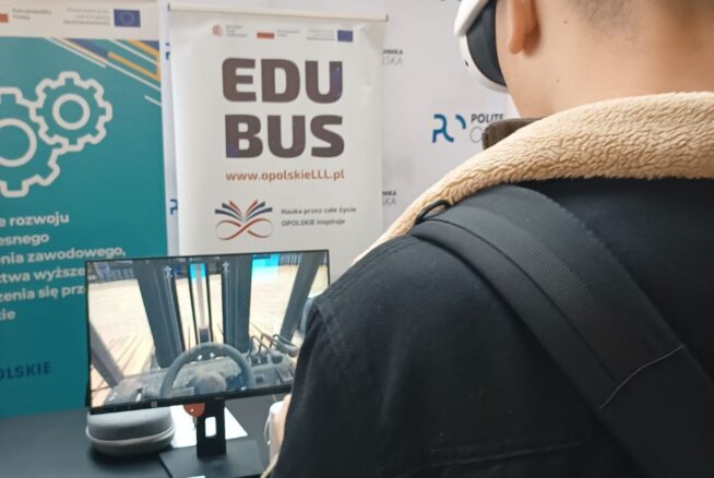 Ujęcie od tyłu, młodego ucznia z założonymi goglami VR. Uczeń skierowany jest w stronę ekranu, na którym wyświetla się kadr z aplikacji „Widzę siebie w tym zawodzie”. W tle, za ekranem znajduje się banner promujący projekt "Wsparcie rozwoju nowoczesnego kształcenia zawodowego..." oraz banner promujący „EduBusa – mobilny punkt szerzenia idei Lifelong Learning” na Opolszczyźnie