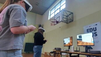 Na zdjęciu widać dwie młode uczennice szkoły podstawowej w sali gimnastycznej, korzystające z gogli VR. Stoją w środku pomieszczenia, wykonując ruchy kontrolerami. W tle znajdują się dwa monitory prezentujące widok z gogli VR oraz roll-up projektu, promujący „EduBusa – mobilny punkt szerzenia idei Lifelong Learning” na Opolszczyźnie. Nad nimi widoczny jest kosz do koszykówki i wysokie okna oświetlające salę.