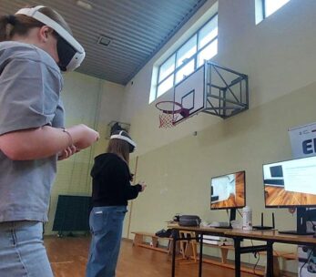 Na zdjęciu widać dwie młode uczennice szkoły podstawowej w sali gimnastycznej, korzystające z gogli VR. Stoją w środku pomieszczenia, wykonując ruchy kontrolerami. W tle znajdują się dwa monitory prezentujące widok z gogli VR oraz roll-up projektu, promujący „EduBusa – mobilny punkt szerzenia idei Lifelong Learning” na Opolszczyźnie. Nad nimi widoczny jest kosz do koszykówki i wysokie okna oświetlające salę.