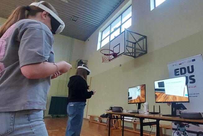 Na zdjęciu widać dwie młode uczennice szkoły podstawowej w sali gimnastycznej, korzystające z gogli VR. Stoją w środku pomieszczenia, wykonując ruchy kontrolerami. W tle znajdują się dwa monitory prezentujące widok z gogli VR oraz roll-up projektu, promujący „EduBusa – mobilny punkt szerzenia idei Lifelong Learning” na Opolszczyźnie. Nad nimi widoczny jest kosz do koszykówki i wysokie okna oświetlające salę.