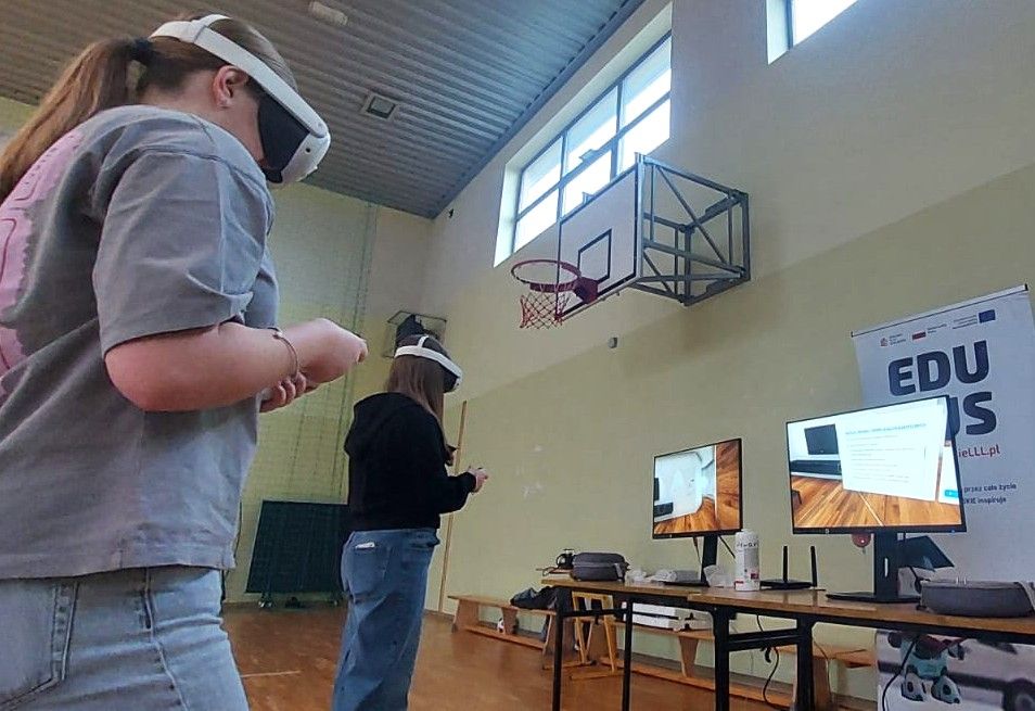 Na zdjęciu widać dwie młode uczennice szkoły podstawowej w sali gimnastycznej, korzystające z gogli VR. Stoją w środku pomieszczenia, wykonując ruchy kontrolerami. W tle znajdują się dwa monitory prezentujące widok z gogli VR oraz roll-up projektu, promujący „EduBusa – mobilny punkt szerzenia idei Lifelong Learning” na Opolszczyźnie. Nad nimi widoczny jest kosz do koszykówki i wysokie okna oświetlające salę.