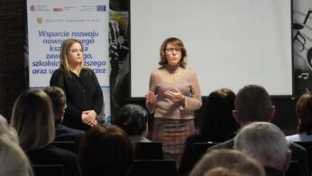 Dwie kobiety stoją na scenie, przemawiając do publiczności, na tle dużego, czystego ekranu projekcyjnego. Obok nich znajduje się banner promujący projekt Starostwa Powiatowego w Nysie dotyczący rozwoju nowoczesnego kształcenia zawodowego. Jest to moment otwarcia lub podsumowania konferencji.