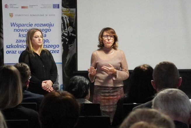Dwie kobiety stoją na scenie, przemawiając do publiczności, na tle dużego, czystego ekranu projekcyjnego. Obok nich znajduje się banner promujący projekt Starostwa Powiatowego w Nysie dotyczący rozwoju nowoczesnego kształcenia zawodowego. Jest to moment otwarcia lub podsumowania konferencji.