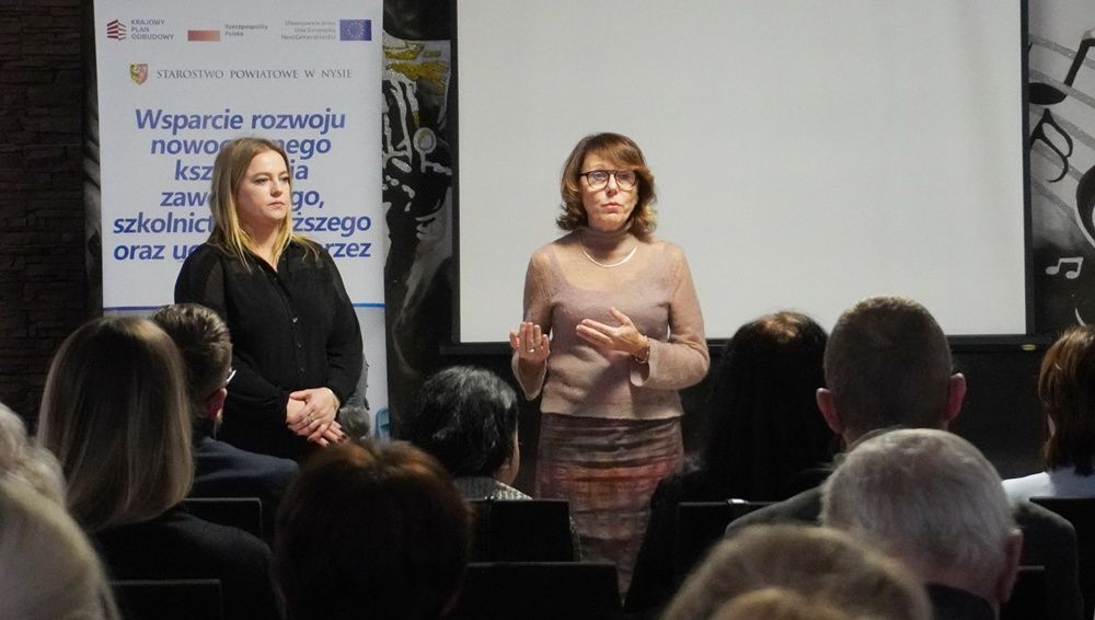 Dwie kobiety stoją na scenie, przemawiając do publiczności, na tle dużego, czystego ekranu projekcyjnego. Obok nich znajduje się banner promujący projekt Starostwa Powiatowego w Nysie dotyczący rozwoju nowoczesnego kształcenia zawodowego. Jest to moment otwarcia lub podsumowania konferencji.
