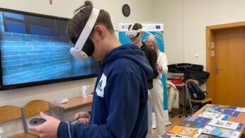 Młody chłopak w ciemnej bluzie z kapturem i goglami VR stoi z kontrolerem w ręce, uczestnicząc w symulacji wirtualnej rzeczywistości. Obok niego, nieco z tyłu, inna uczennica również ma założone gogle VR, a dalej widać instruktorkę. Na dużym ekranie z tyłu wyświetla się ściana z teksturą cegieł, a na ławkach leżą materiały edukacyjne.