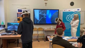 Widok na salę lekcyjną, w której uczniowie aktywnie korzystają z gogli wirtualnej rzeczywistości (VR) podczas zajęć. Na dwóch dużych monitorach widoczne są różne symulacje, w tym jazda samochodem i elementy konstrukcyjne. W tle znajduje się baner informujący o wsparciu na rzecz nowoczesnego kształcenia, a zajęcia prowadzone są w dynamicznej atmosferze.