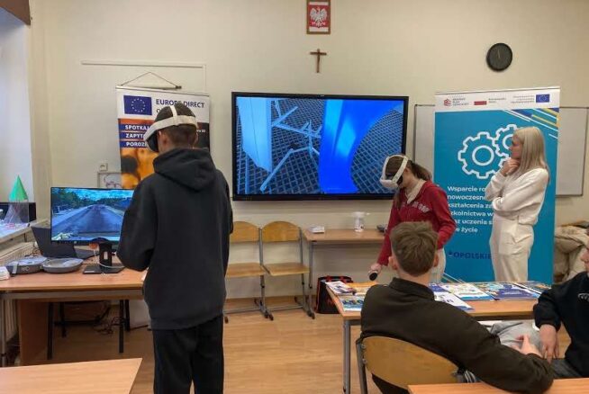 Widok na salę lekcyjną, w której uczniowie aktywnie korzystają z gogli wirtualnej rzeczywistości (VR) podczas zajęć. Na dwóch dużych monitorach widoczne są różne symulacje, w tym jazda samochodem i elementy konstrukcyjne. W tle znajduje się baner informujący o wsparciu na rzecz nowoczesnego kształcenia, a zajęcia prowadzone są w dynamicznej atmosferze.